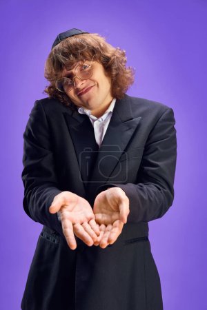 Photo for Portrait of young Jewish man with long hair and in glasses, in kippa holds hands near camera against purple background. Purim, festival, holiday, celebration Pesach or Passover, Judaism concept. Ad - Royalty Free Image