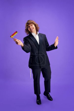 Photo for Joyful man in suit holding grogger symbol used to drown out name of Haman villainous character against purple backdrop. Celebration of Purim, festival, holiday, Judaism, religion concept. Ad - Royalty Free Image