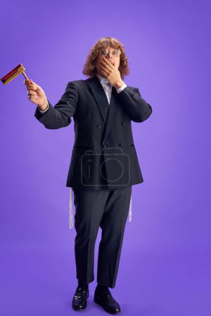 Photo for Full length portrait of young scared Jewish man holding grogger symbol used to drown out name of Haman villainous character. Purim, business, festival, holiday, celebration, religion concept. - Royalty Free Image
