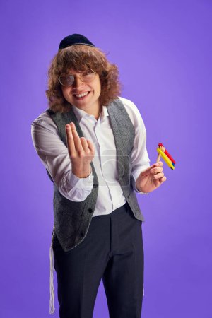 Photo for Young joyful Jewish man in kippah and waistcoat holds grogger and implying inspiration against purple background. Purim, business, festival, holiday, celebration Pesach or Passover concept. - Royalty Free Image