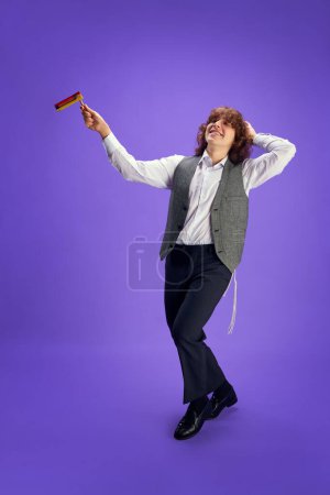 Retrato de joven judío feliz en traje formal y kippah bailando con grogger sobre fondo púrpura estudio. Purim, negocio, festival, día de fiesta, celebración, judaísmo, concepto de religión.