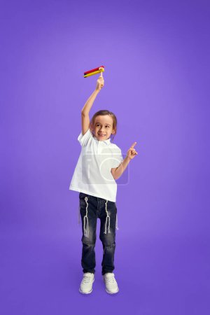 Foto de Niño feliz en traje casual sosteniendo juguete grogger colorido y mostrando gesto apuntando contra fondo de estudio púrpura. Purim, negocio, festival, día de fiesta, celebración Pesaj o concepto de Pascua. - Imagen libre de derechos