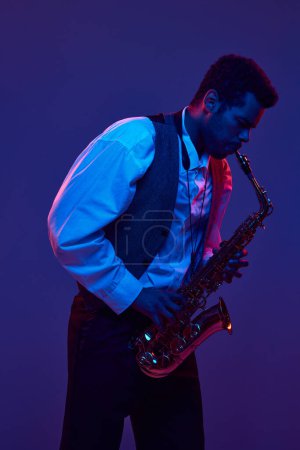 Téléchargez les photos : Homme afro-américain en chemise et gilet jouant du saxophone sur fond bleu et violet dans l'éclairage au néon. Concept de jazz, blues, musique instrumentale chic, festivals et concerts. Publicité - en image libre de droit
