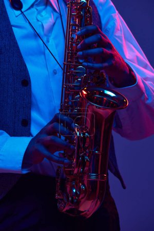 Téléchargez les photos : Photo recadrée d'un homme de jazz afro-américain jouant du saxophone, instrument à vent sur fond dégradé dans une lumière au néon mixte. Concept d'instrument de musique classique, concerts et festivals. Publicité - en image libre de droit