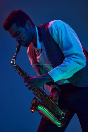 Téléchargez les photos : Virtuose afro-américain, musicien solo jouant des mélodies de jazz sur saxophone sur fond bleu dégradé sous néon mixte. Concept de jazz, blues, musique instrumentale chic, concert. Publicité - en image libre de droit