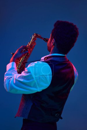Téléchargez les photos : Vue arrière de l'homme jouant du saxophone jouant des mélodies de jazz sur fond bleu vif dans un fond mixte rose-violet. Concept de jazz, blues, musique instrumentale chic, festivals et concerts. Publicité - en image libre de droit