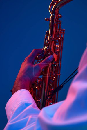 Téléchargez les photos : Portrait recadré de jeune homme tenant du saxophone et jouant des mélodies blues sur fond bleu au néon. Des accords. Concept d'instrument de musique classique, concerts et festivals. Publicité - en image libre de droit