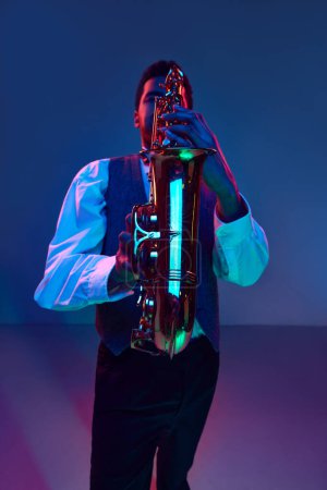 Téléchargez les photos : Jazzman. Élégant saxophoniste habillé chemise blanche et gilet jouant des mélodies de jazz dans la lumière bleu-rose sur fond de studio dégradé. Concept de jazz, blues, musique instrumentale chic, festivals. Publicité - en image libre de droit