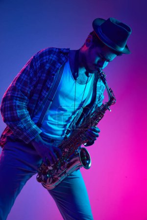 Téléchargez les photos : Homme afro-américain habillé rétro, en chapeau jouant du saxophone sur fond rose dégradé et bleu néon. Concept de jazz, blues, musique instrumentale chic, festivals et concerts. Publicité - en image libre de droit