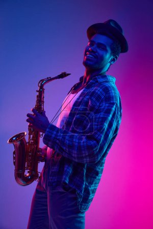 Téléchargez les photos : Portrait mi-long de l'homme afro-américain, saxophoniste jouant de la musique jazz sur fond dégradé violet-rose. Concept de jazz, blues, musique instrumentale chic. Publicité - en image libre de droit