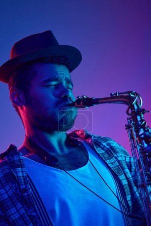 Téléchargez les photos : Portrait d'homme afro-américain, musicien concentré jouant du saxophone en chapeau sur fond bleu et rose dans l'éclairage au néon. Concept de jazz, blues, musique instrumentale chic, festival, concert. Publicité - en image libre de droit