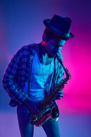 Téléchargez les photos : Musicien intense, homme afro-américain au chapeau jouant du saxophone dans un éclairage vif sur fond rose-bleu. Concept de jazz, blues, musique instrumentale chic, festivals et concerts. Publicité - en image libre de droit