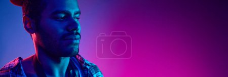 Téléchargez les photos : Portrait de jazzman afro-américain en chapeau regardant loin dans la lumière néon sur fond bleu-rose dégradé avec un espace négatif pour insérer du texte. Concept de jazz, blues, musique instrumentale chic. Publicité - en image libre de droit