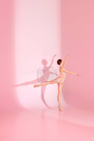 Photo for Ballet in Blush. Graceful dancer, dressed in pink swimsuit, poses barefoot against pastel pink background. Her shadow mirrors her elegance. Concept of poise, beauty, ballet, gracefulness. - Royalty Free Image