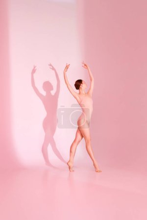 Photo for Pastel Ballet. Woman gracefully poses in pink swimsuit against soft pink backdrop. Her barefoot silhouette casts shadow, echoing her elegant movements. Concept of ballet, grace, beauty. - Royalty Free Image