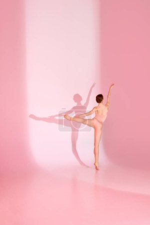 Photo for Pink Elegance. Woman in sporty swimsuit dancing elegantly against pastel studio background. Shadow captures her grace. Concept of ballet and sport, beauty, elegance, movement, shadowplay. - Royalty Free Image
