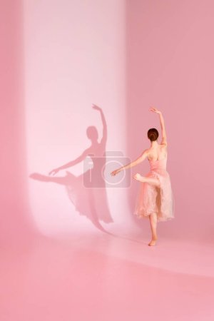 Photo for Poised Elegance. Ballerina in pink dress, tutu poses barefoot against pastel pink background with her shadow. Concept of art, grace, ballet, beauty and femininity. - Royalty Free Image