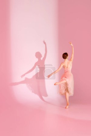 Photo for Balletic Beauty. Classic ballerina dances barefoot in pink dress against pastel pink background with her shadow. Concept of art and hobby, elegance, ballet, beauty and femininity. - Royalty Free Image