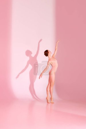 Photo for Pink Elegance. Woman in sporty swimsuit dancing elegantly against pastel studio background. Shadow captures her grace. Concept of ballet, beauty, elegance, movement, shadowplay. - Royalty Free Image