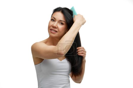 Beautiful middle aged woman combing her healthy and silky long hair looking away against white studio background. Concept of hair care, health, beauty treatment, care and spa procedures.