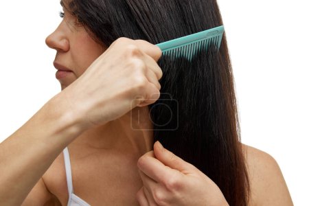 Close up photo of beautiful middle aged woman brushing her healthy and silky long hair looking away against white studio background. Concept of hair care, beauty treatment, care and spa procedures.