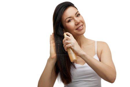 Beautiful middle aged woman sprays hair styling product on her long and thick silky hair against white studio background. Concept of hair care, health, beauty treatment, care and spa procedures.
