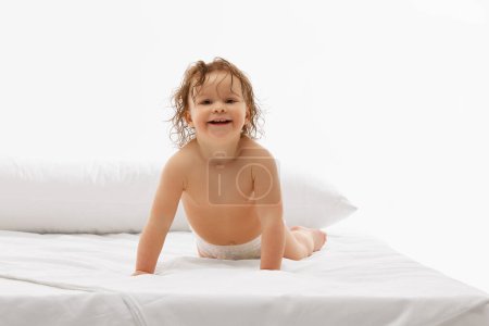 Little girl, 2 ears old baby with lively expression, experiencing comfort of plush white mattress against white background. Concept of childhood, motherhood, life, birth. Copy space for ad
