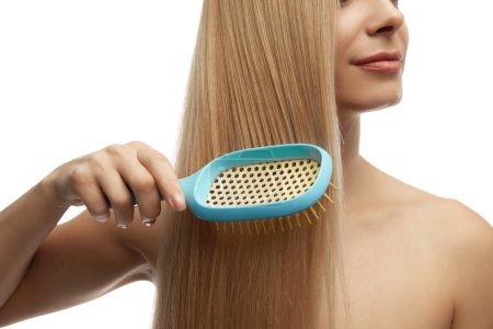 Close up photo of woman combing blonde, healthy and silky hair against white studio background. Beauty and health of hair. Concept of natural beauty, cosmetology, female health, hair care. Ad