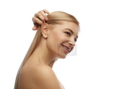 Side profile view photo of smiling woman holding her ponytail against white studio background. Hairstyles and hair care treatments. Concept of natural beauty, anti aging, cosmetology, female health.
