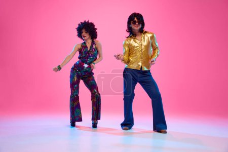 Man and woman dressed in vibrant 1970s attire dancing in motion against gradient pink studio background. Retro fashion. Concept of American culture, 1970s, 1980s fashion, music, comparisons of eras.