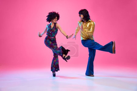 Mann und Frau in pulsierendem 70er-Jahre-Gewand tanzen vor rosa Studiohintergrund in Bewegung. Retro-Mode. Konzept der amerikanischen Kultur, Mode der 1970er, 1980er Jahre, Musik, Epochenvergleiche.