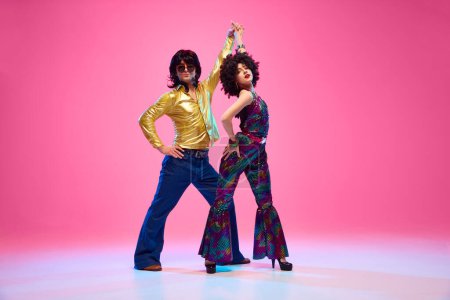 Groovy dance couple in bright attire performing disco moves in motion against gradient pink studio background. Concept of American culture, 1970s, 1980s fashion, music, comparisons of eras.