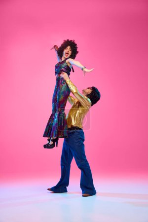 To rhythm of dance. Talented couple, dance partners in dynamic disco pose against gradient pink studio background.Concept of American culture, 1970s, 1980s fashion, music, comparisons of eras.