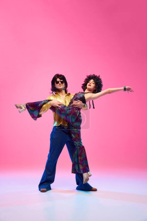 Dynamisches Porträt eines Tänzerduos, Mann und Frau, das in Retro-Kleidung vor gradienten rosa Studiohintergrund posiert. Konzept der amerikanischen Kultur, Mode der 1970er, 1980er Jahre, Musik, Epochenvergleiche.