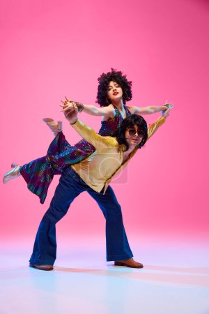Disco tanzendes Paar in dynamischer Hebebühne posiert im lebhaften Retro-Outfit vor gradienten rosafarbenen Studiohintergrund. Energetische Bewegungen. Konzept der amerikanischen Kultur, 1970er Jahre, Musik, Epochenvergleiche.