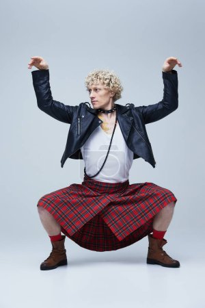 Foto de Hombre en escocés y chaqueta de cuero posando con los brazos levantados, encarnando una mezcla de estilo tradicional y punk sobre fondo gris estudio. Concepto de fusión de moda, arte, singularidad, auto-expresión. Anuncio - Imagen libre de derechos