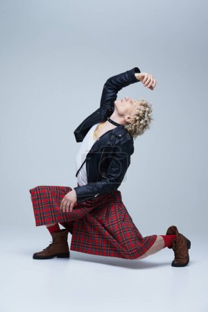 Foto de Hombre en escocés y chaqueta de cuero bailando con los brazos levantados, encarnando mezcla de estilo tradicional y punk sobre fondo gris estudio. Concepto de fusión de moda, arte, singularidad, auto-expresión. Anuncio - Imagen libre de derechos