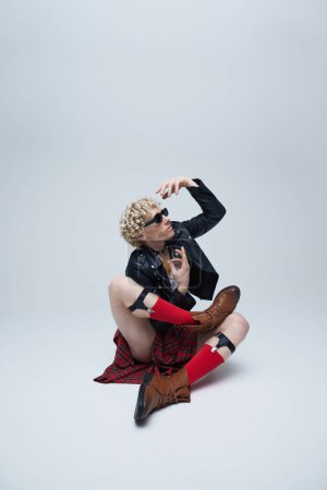 Foto de Elegante bailarín masculino que combina elementos punk y atuendo escocés en movimiento enérgico contra fondo gris del estudio. Concepto de fusión de moda, fotografía de arte, estilo, singularidad, auto-expresión. Anuncio - Imagen libre de derechos