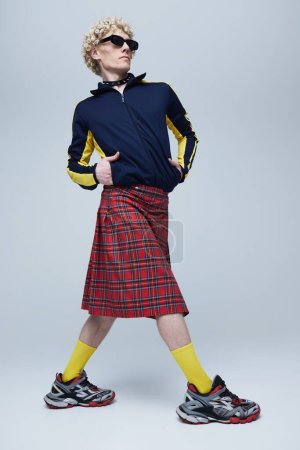 Foto de Retrato de cuerpo entero del joven hombre de pelo rizado posando en escocés escocés y ropa deportiva sobre fondo gris estudio. Concepto de fusión de moda, estilo, queer, singularidad, auto-expresión. Anuncio - Imagen libre de derechos