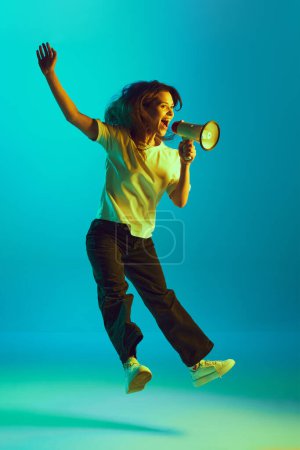 Ganztägiges Porträt einer jungen Frau, die in gelbem Neonlicht vor dem Studiohintergrund wichtige Neuigkeiten ins Megafon ruft. Konzept der sozialen Medien, Kommunikation, Nachrichten, Botschaften.