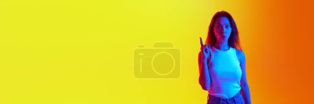 Banner. Portrait of female fashion model pointing up in neon light against yellow-orange background with negative space to insert text. Concept of human emotions, self-expression, beauty, youth. Ad