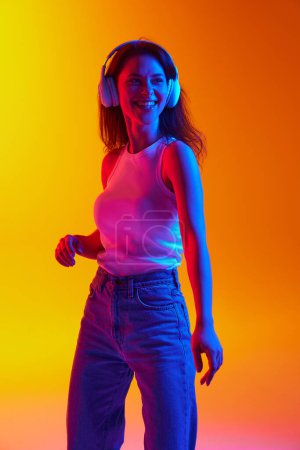 Portrait of young lady dressed casual outfit listening music in headphones against gradient studio background in neon light. Concept of human emotions, youth culture, self-expression, technology. Ad