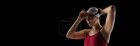 Banner. Athletic of young female swimmer wearing cap and goggles before swim race against black studio background. Negative space. Concept of professional sport, motion, strength and power. Ad