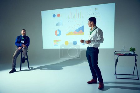 Photo for Two men present at business meeting, one seated listening, other standing, gesturing to data chart in conference room. Concept of business, startup, leadership and personal development courses. - Royalty Free Image