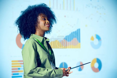 Téléchargez les photos : Femme afro-américaine présentant des données statistiques avec pointeur sur fond de graphique projeté lors d'une conférence d'affaires. Concept d'entreprise, de démarrage, de leadership et de développement personnel. - en image libre de droit