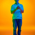 Full-length portrait of smiling Asian man dressed casual outfit messaging in vibrant neon light against gradient orange background. Concept of human emotions, fashion and beauty, work and hobby.