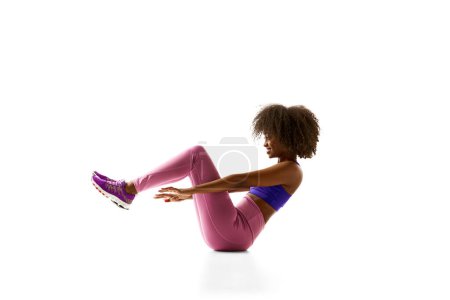 Athletische Afroamerikanerin in lila Aktivkleidung balanciert in V-Sit-Position vor weißem Studiohintergrund. Konzept von Sport, Trauerroutine, aktiver und gesunder Lebensweise, Energie, Aktion.