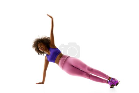 Femme athlétique afro-américaine en équilibre de vêtements de sport violet, s'entraînant sur fond de studio blanc. Concept de sport, routine de deuil, mode de vie actif et sain, énergie, action.