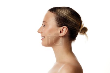 Profile photo of young beautiful woman with bare shoulders smiling against white studio background. Double chin prevention. Concept of natural beauty people, injections, cosmetology. Ad