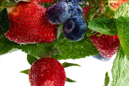 Nahaufnahme von frischen Erdbeeren und Blaubeeren in Wasser auf grünen Minzblättern. Textur des kühlenden süßen Sommergetränks mit Makroblasen an der Glaswand. Konzept von Speisen und Getränken, Ernährung, Frische